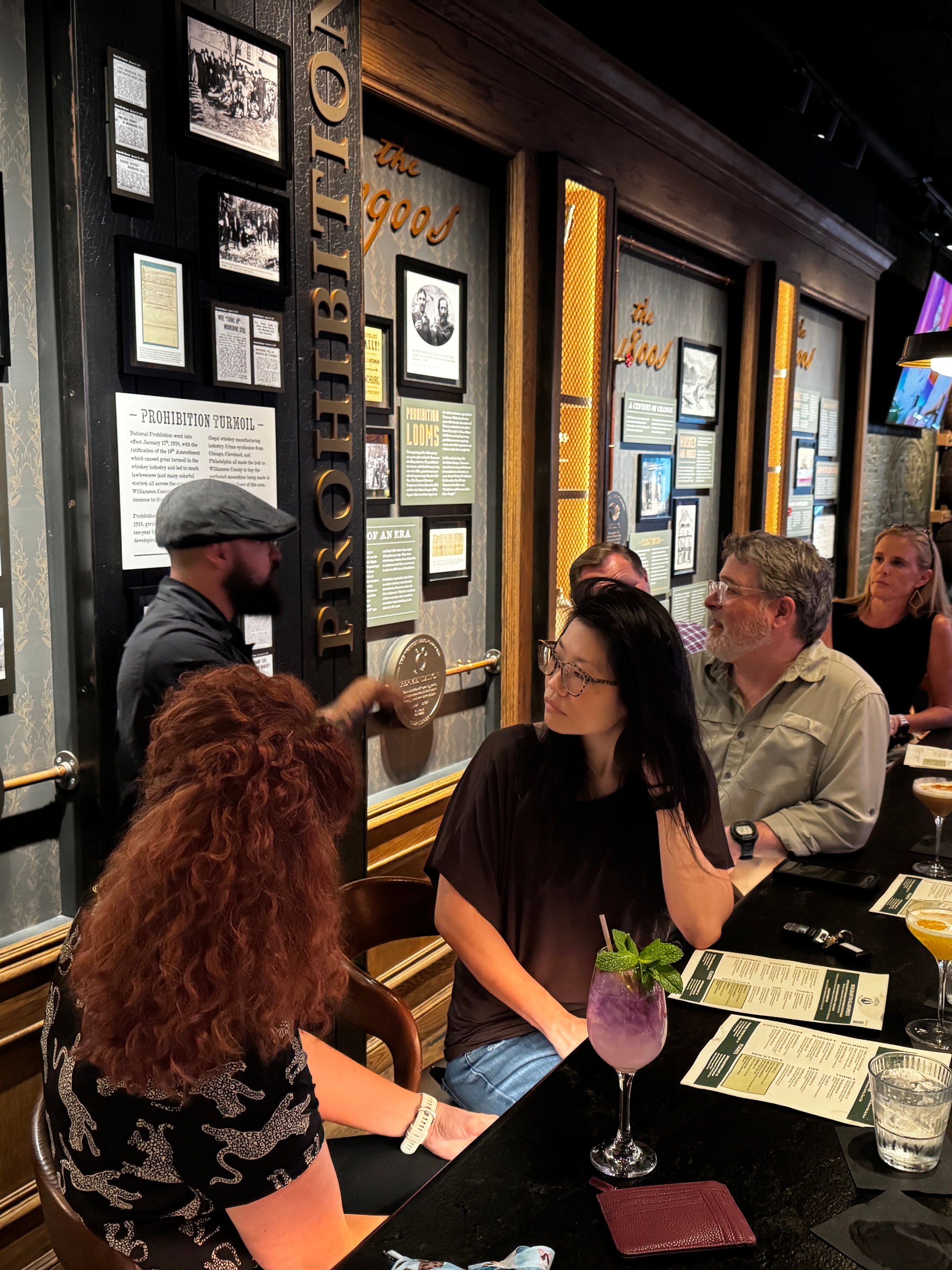 People seated at a bar in a dimly lit space with framed pictures on the wall.
