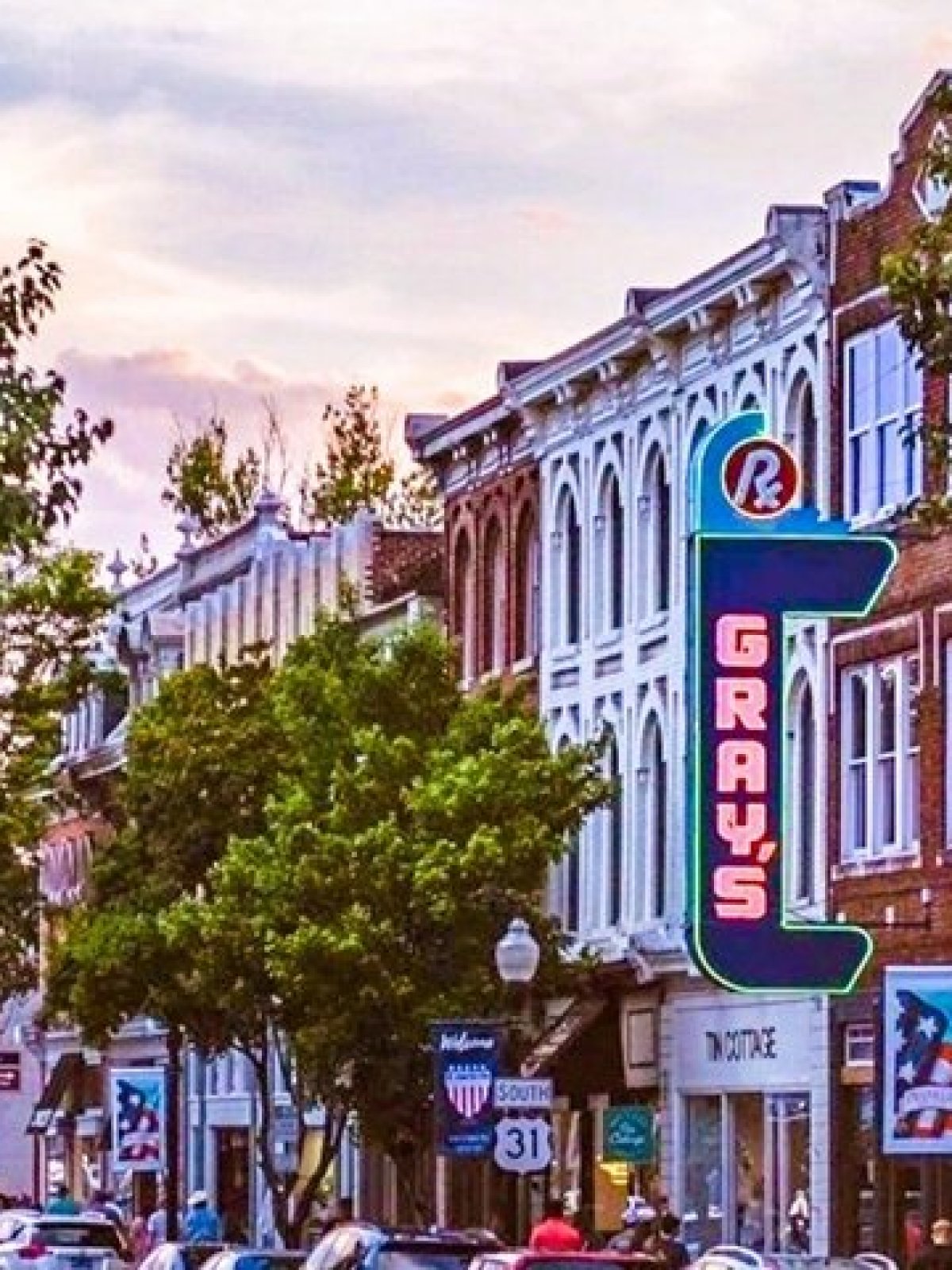 Lively street with historic buildings, trees, and 'Gray's' neon sign at sunset.