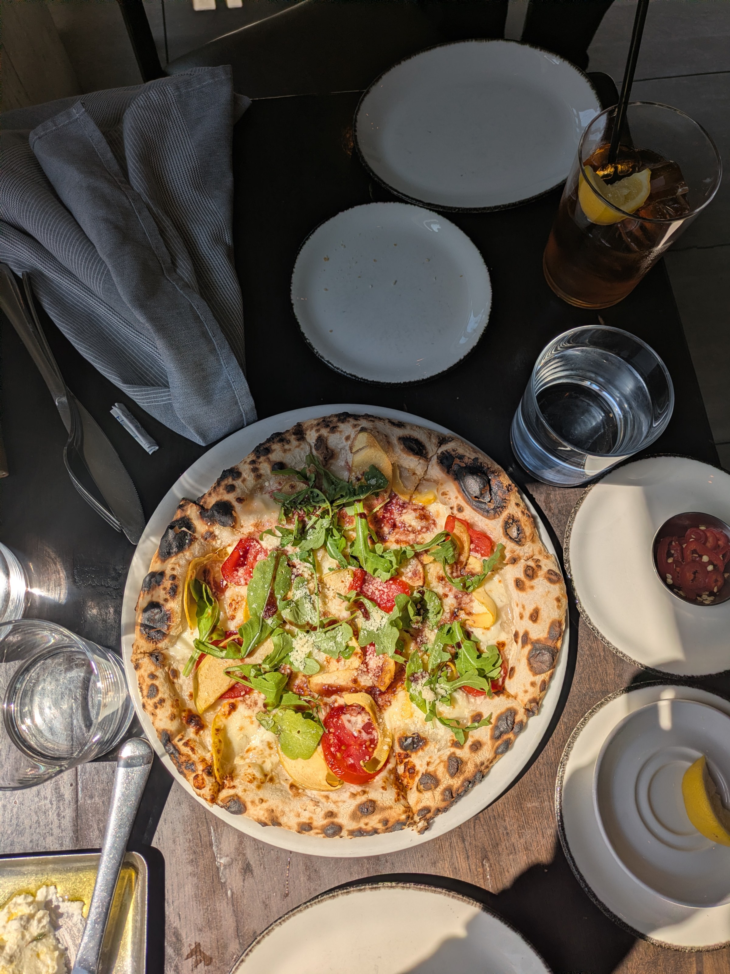 Pizza with tomatoes and greens on a table with plates, drink, and utensils.