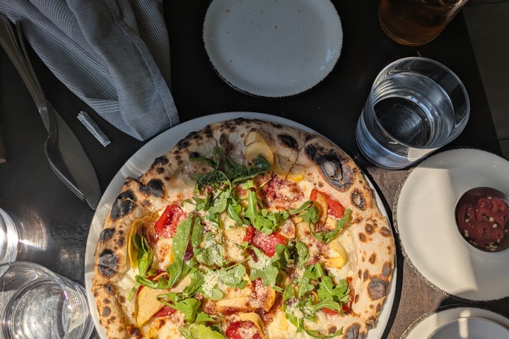 Pizza with tomatoes and greens on a table with plates, drink, and utensils.